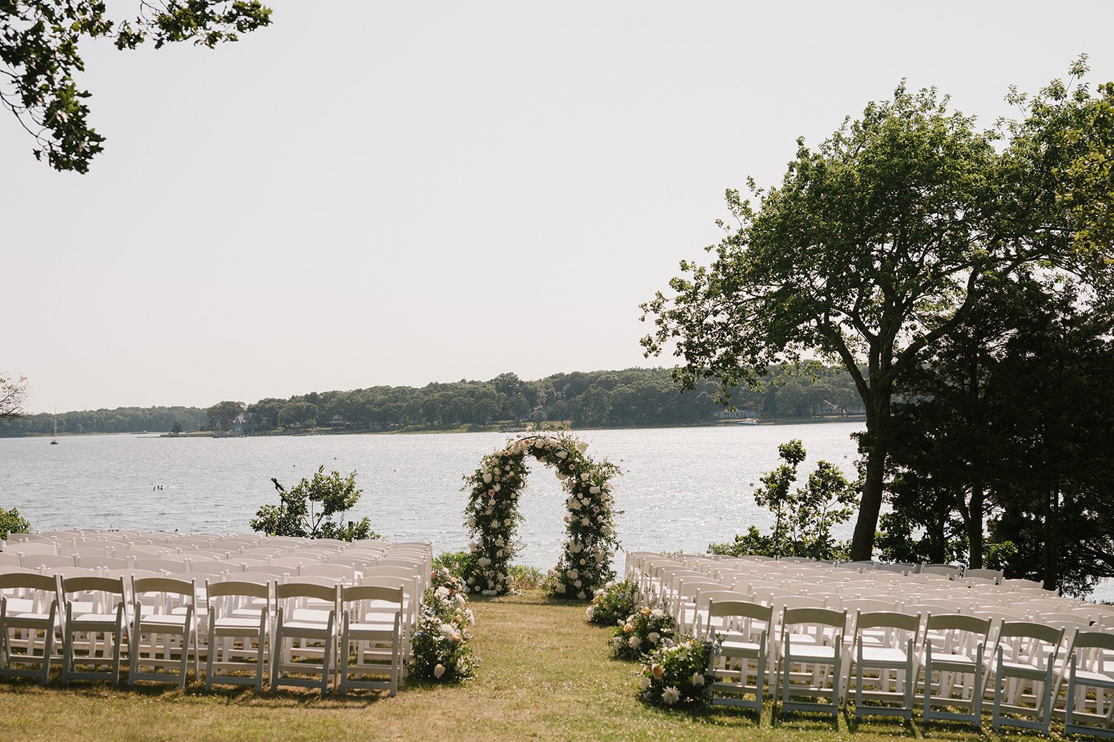 Outdoor wedding setup by a lake with white chairs arranged in rows facing a floral arch, surrounded by greenery and trees.